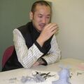 A photograph of Kaneko with prototype versions of the figures he designed for Shin Megami Tensei Card Game: Nocturne Ver. Part 2